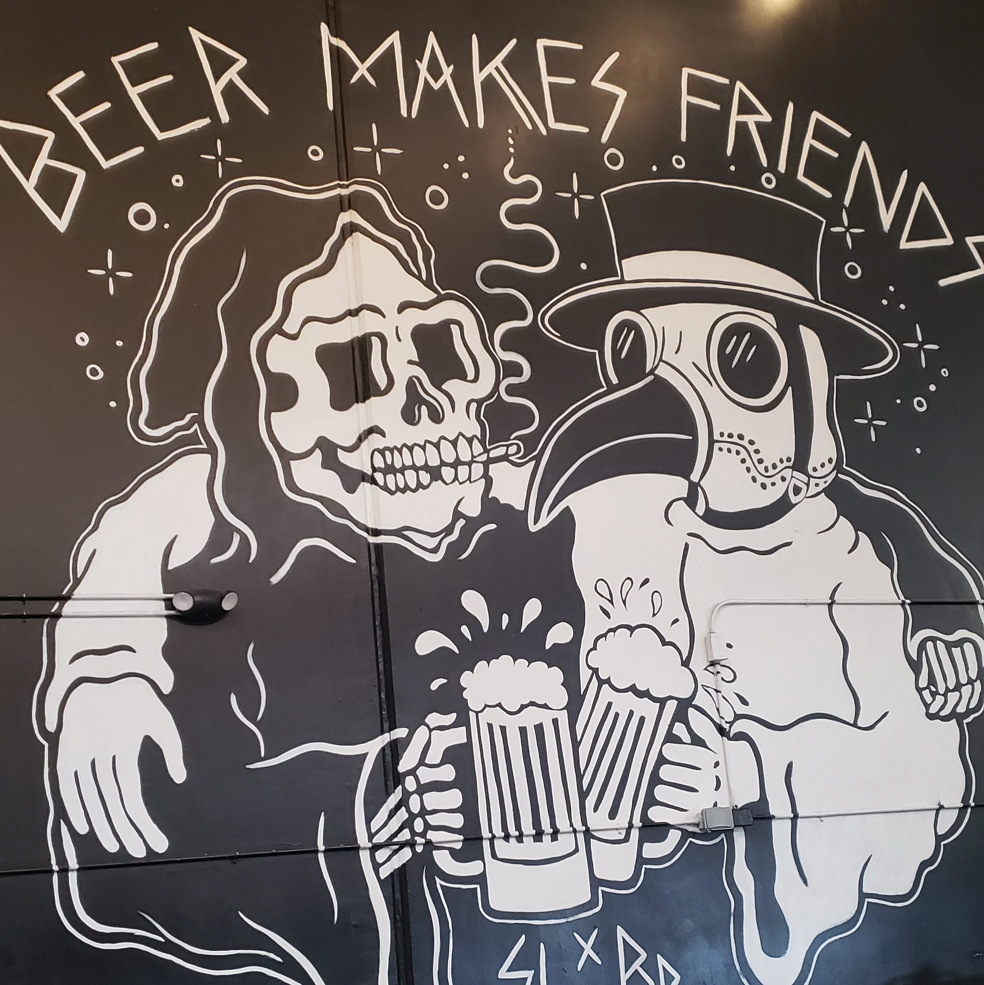 On a wall, a black and white mural painting of the plague doctor and the grim reaper toasting each other with full beers and their arms around each other. The grim reaper is smoking. At the top it says "Beer makes friends."