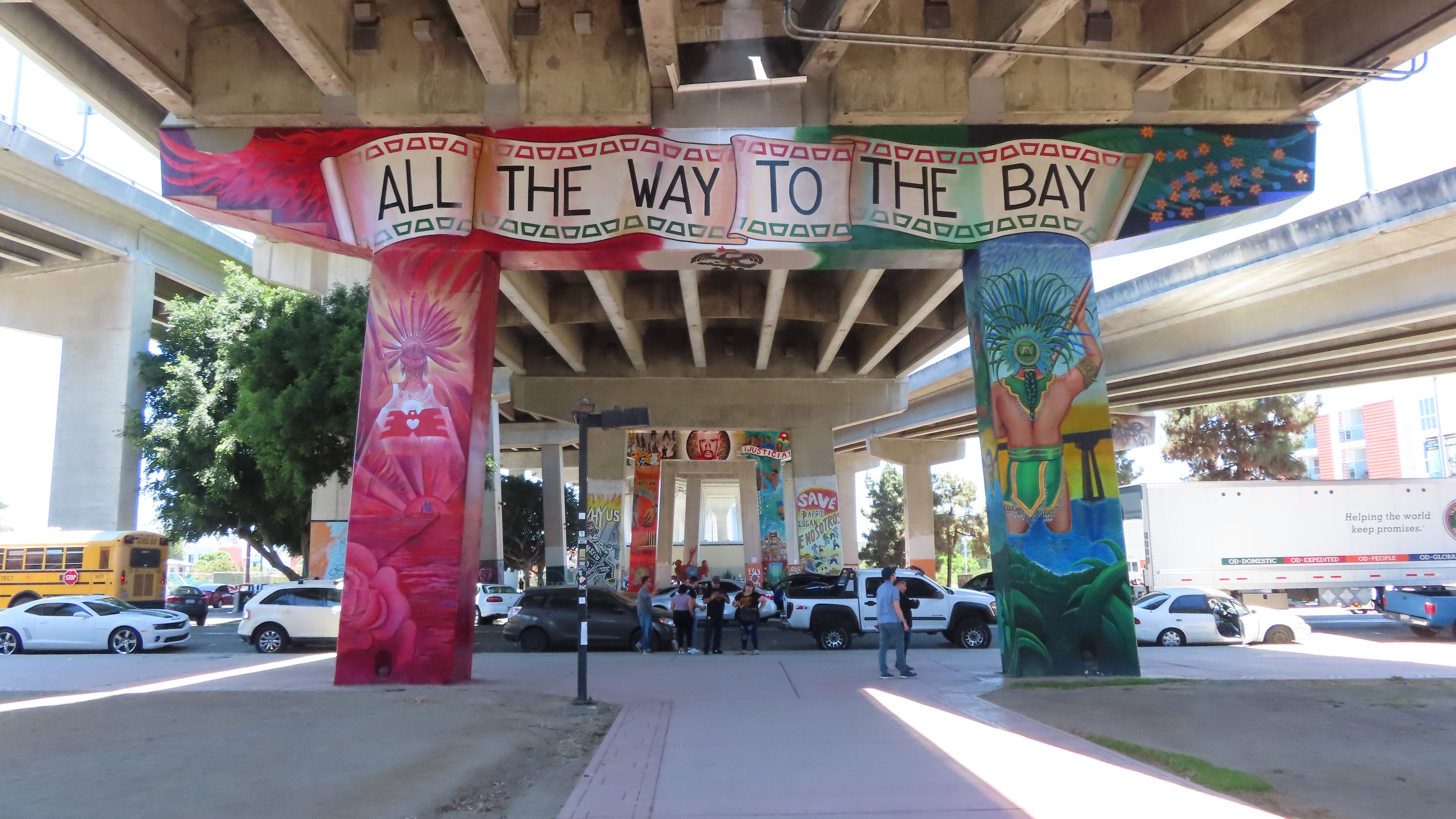 A mural on bridge pylons of the Coronado Bridge in Chicano Park, Barrio Logan. The mural depicts Indigenous gods or warriors overlooking the San Diego Bay and Coronado bridge. One is in more realistic colors and the other pylon depicts the same warrior in shining neon pink. The top says, "All the Way to the Bay" in reference to extending the murals along the bridge pylons to the edge of the San Diego Naval Base along the water.