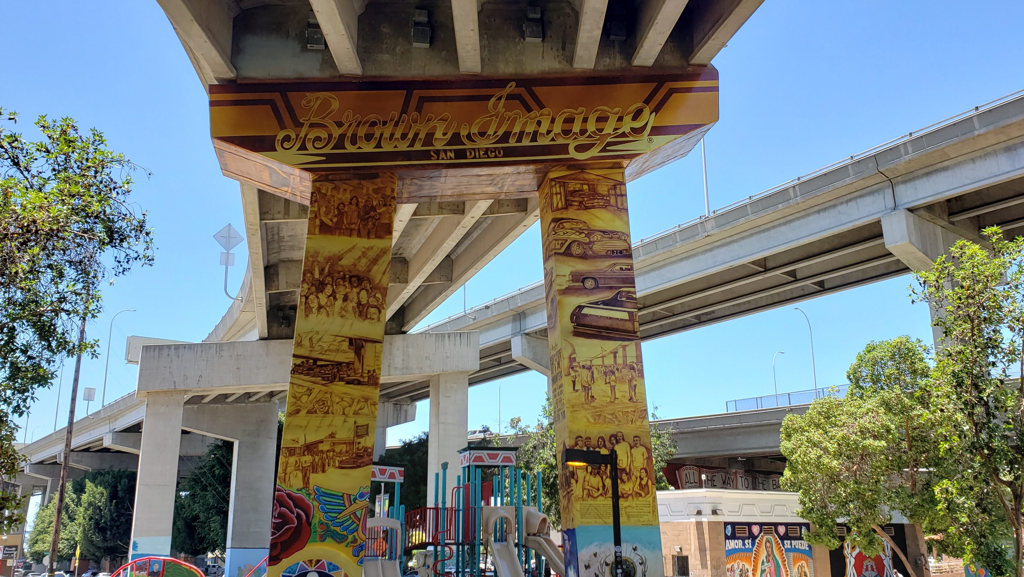 A mural on the pylon bridges supporting a freeway in what is known as Chicano Park. The mural says "Brown Image" at the top showcasing a group of individuals into car culture who rode lowriders and supported the park and its artistic efforts. There is a rose and a bird on the bottom with depictions of men and women at the demonstration site looking at the newly constructed Coronado Bridge in 1970.