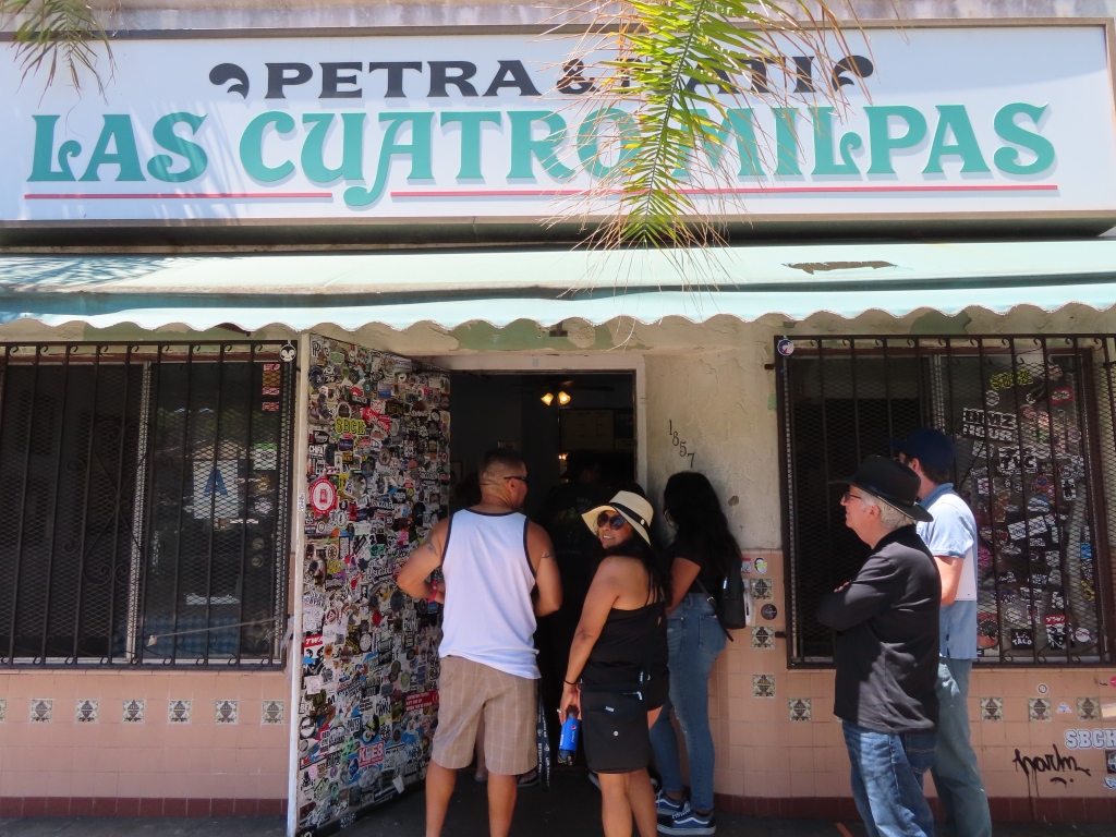 The entrance and above the sign outside the main building of Las Cuatro Milpas. The door and right window are covered in bumper stickers. Above the name of the restaurant is the founders Petra and Nati. There is a woman in a black outfit with a sunhat looking back. Matthew and Bob wait to go inside.