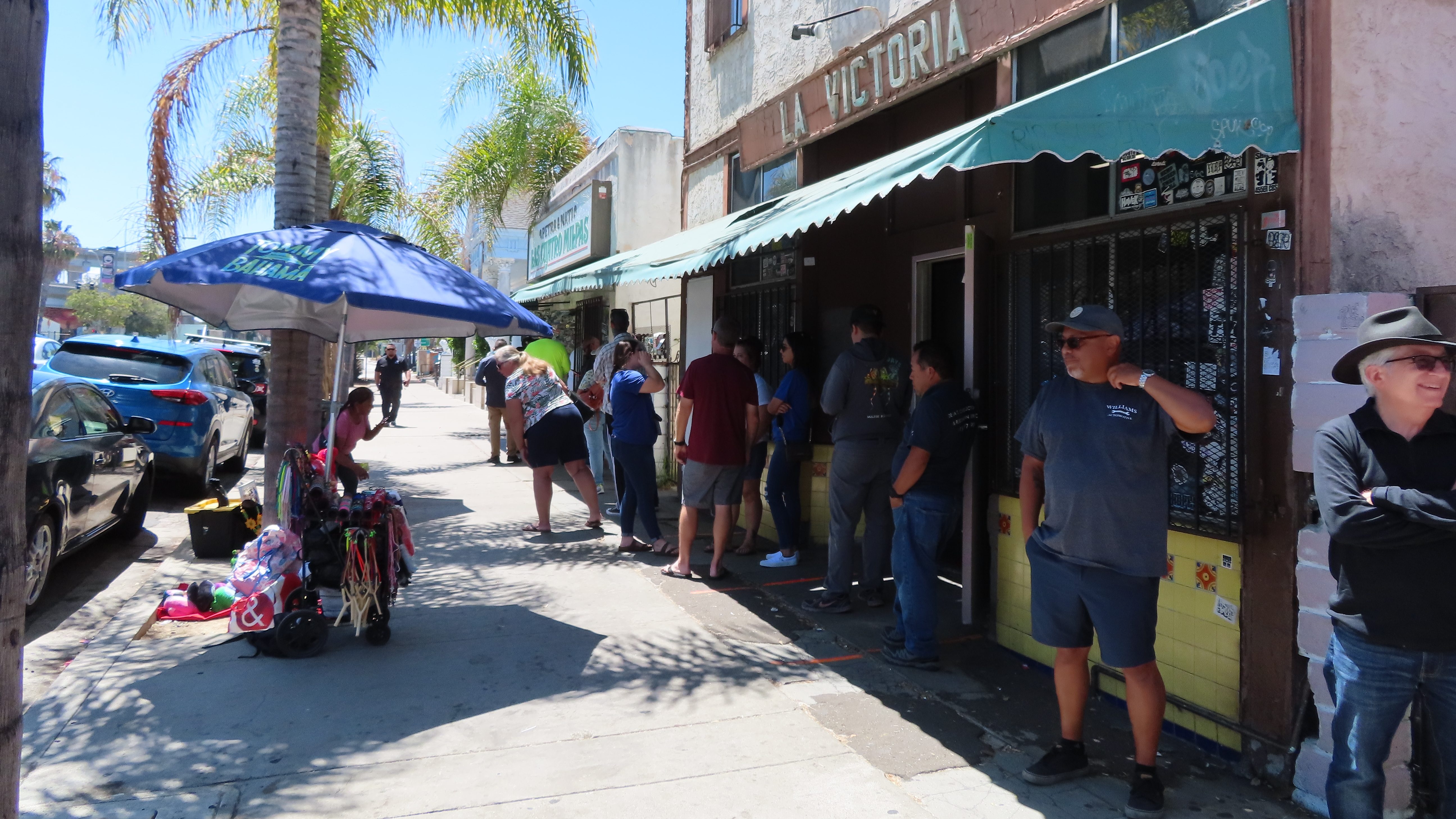 Candid shot of line for food outside Los Cuatro Milpas. There is a guy tugging at his shirt in the heat. A person is looking over at a small stall where a woman sells jewelry and other items to people in line at the restaurant. It stretches for 70-80 feet or so across the length of the restaurant. There are palm trees along the sidewalk.