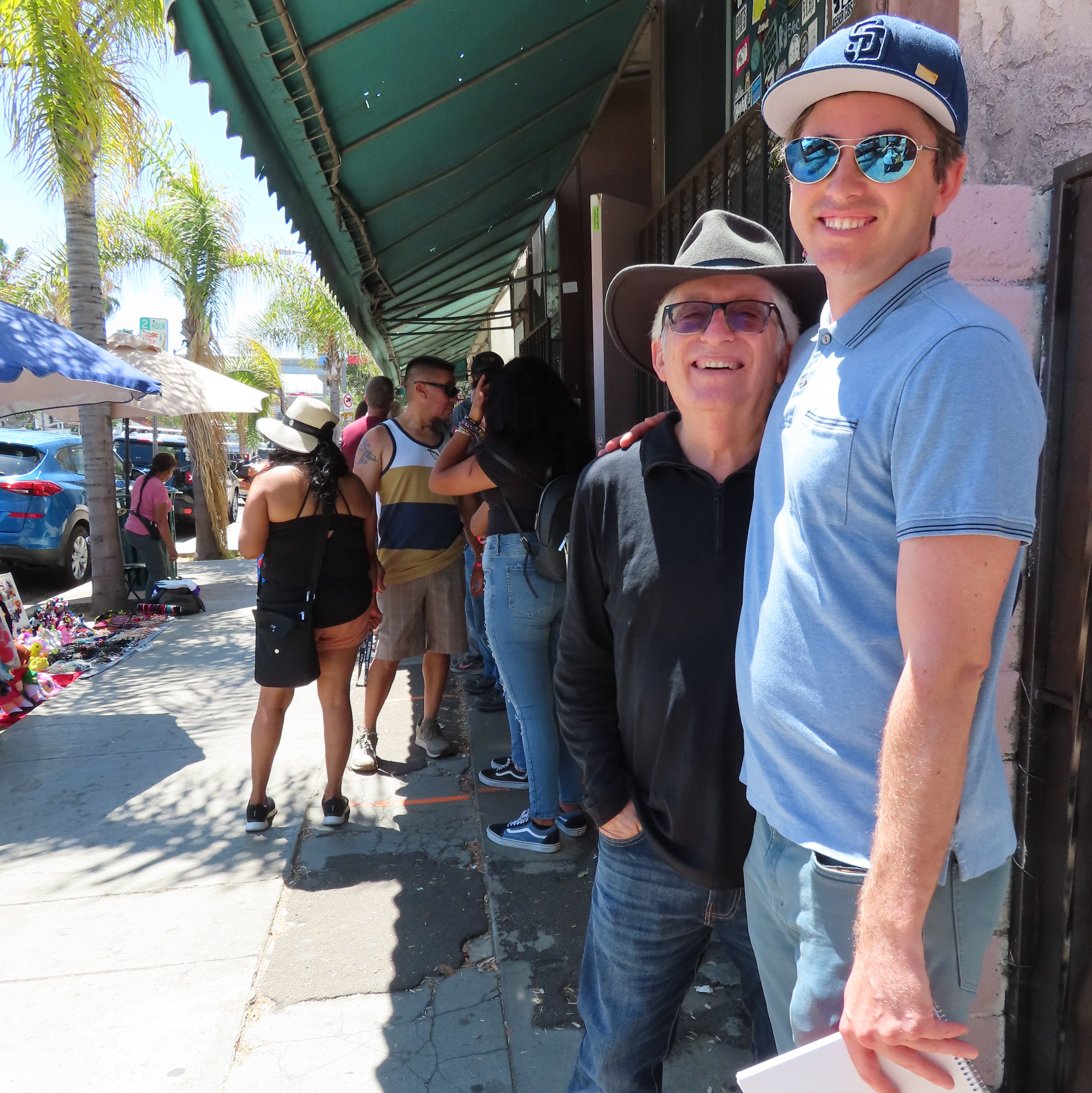 Matthew and Bob, his dad in line at Las Cuatro Milpas. They smile. Matthew has sunglasses and a SD hat on. Bob has jeans, a black shirt and a green sunhat on. There are people milling about, waiting in the background.