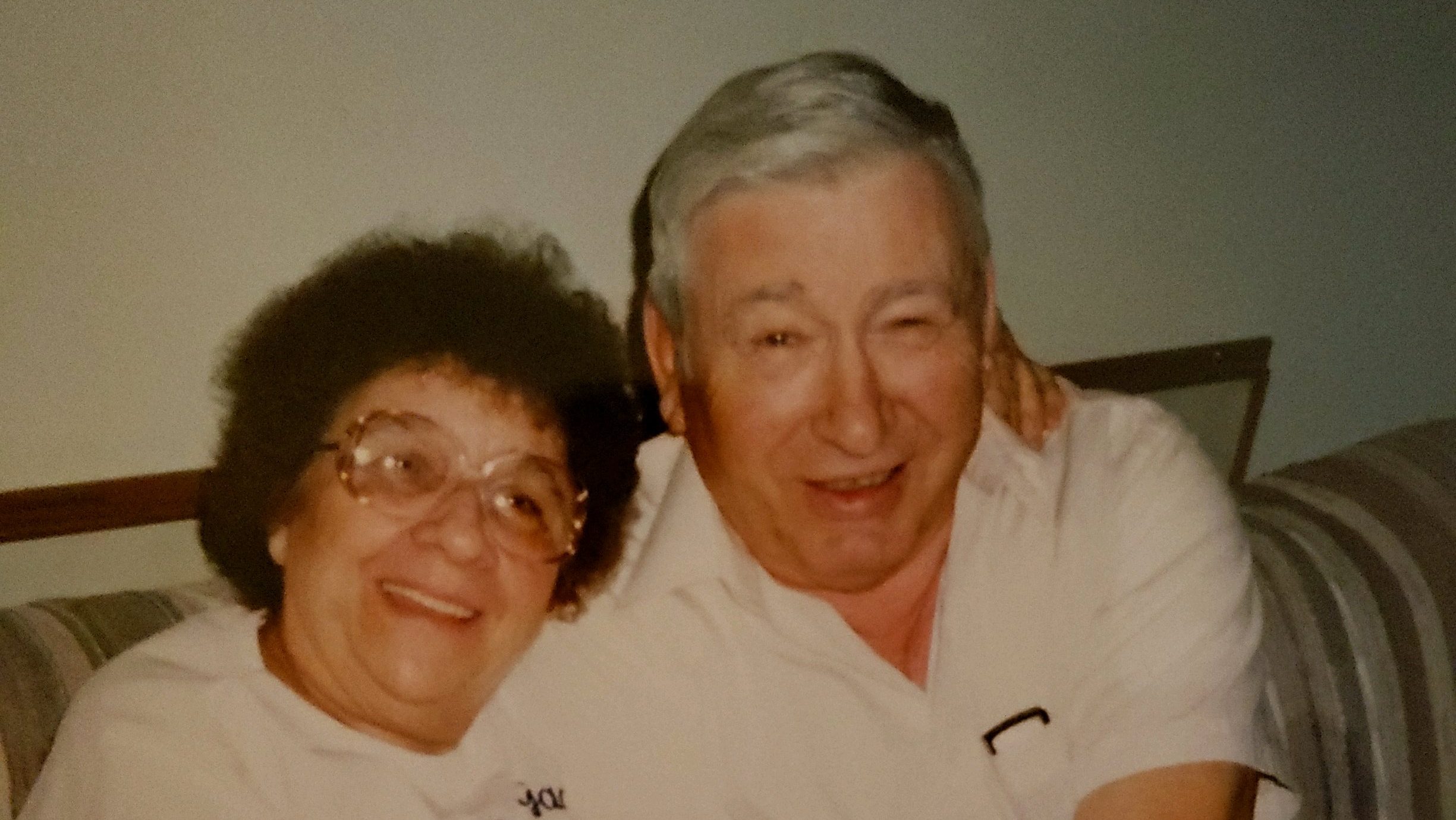 Picture of Florence Manly and Bill Manly in their home on their living room couch. Florence is on the left with fuzzy dark hair, big glasses, puffy eyes and a smile. Bill has squinted eyes, gray hair and glasses in his shirt pocket. They both have on white shirts and are in their 70s.