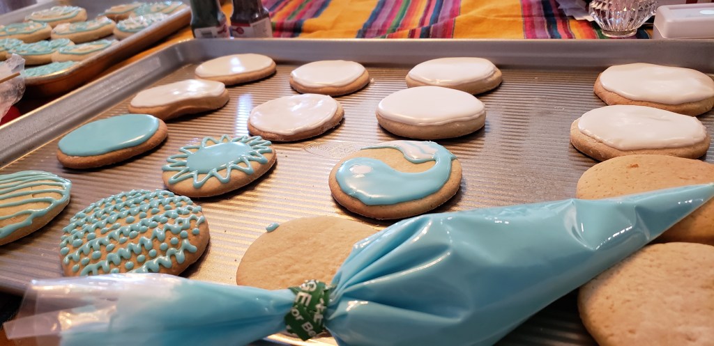 The process of icing the cookies. There is a baking sheet tray on a tablecloth filled with cookies. A piping bag is laying on the uniced cookies with baby blue icing. The cookies toward the back have white icing, the cookies toward the front are decorated with blue icing. There are intricate designs on some of the cookies in front. They are works in progress at this point.