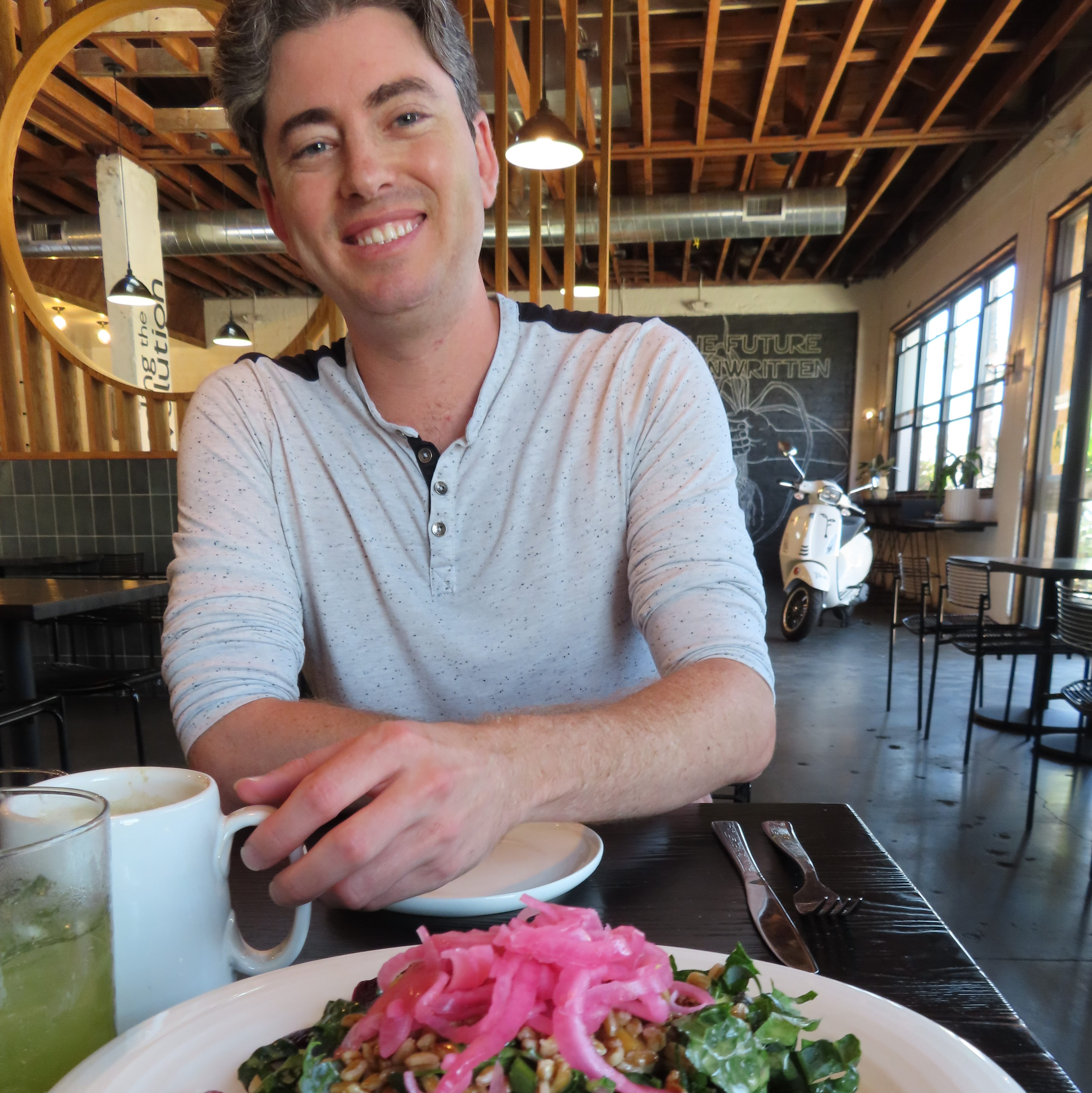 Matthew at The Plot smiling into the camera grabbing his coffee. The motorbike is behind with the propped open entrance door. There are empty tables and chairs behind and across the room is a mural of an arm pulling out a plant root with the words, "The Future is Unwritten." I am dressed in a long sleeve, white shirt.