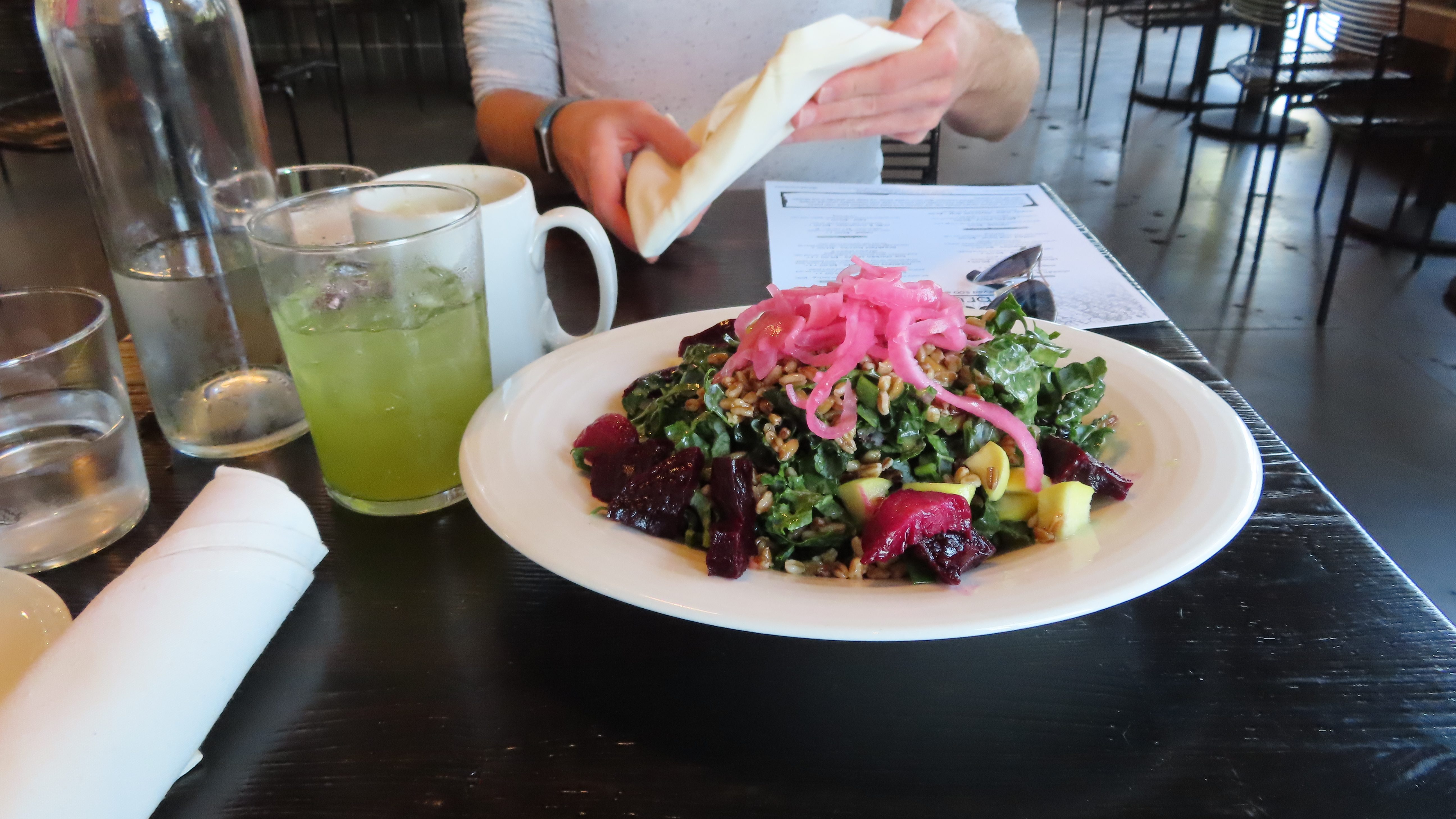 A kale salad with farro, pickled red onion, apples and cara cara rice dressing. The colors of yellow from the apple, red from the pickled onion and green kale looks bright and beautiful. There is my coffee mug and Jennifer's green drink on the right.