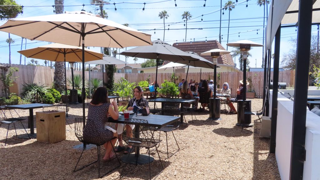 The patio outside The Plot with people eating and enjoying their meal. Tan-colored woodchips cover the area with edible plants bordering the perimeter. There are light beige umbrellas around black tables for shade and heaters for cooler weather at dinner service. There is a brown picket that surrounds the spacious property and blue skies.