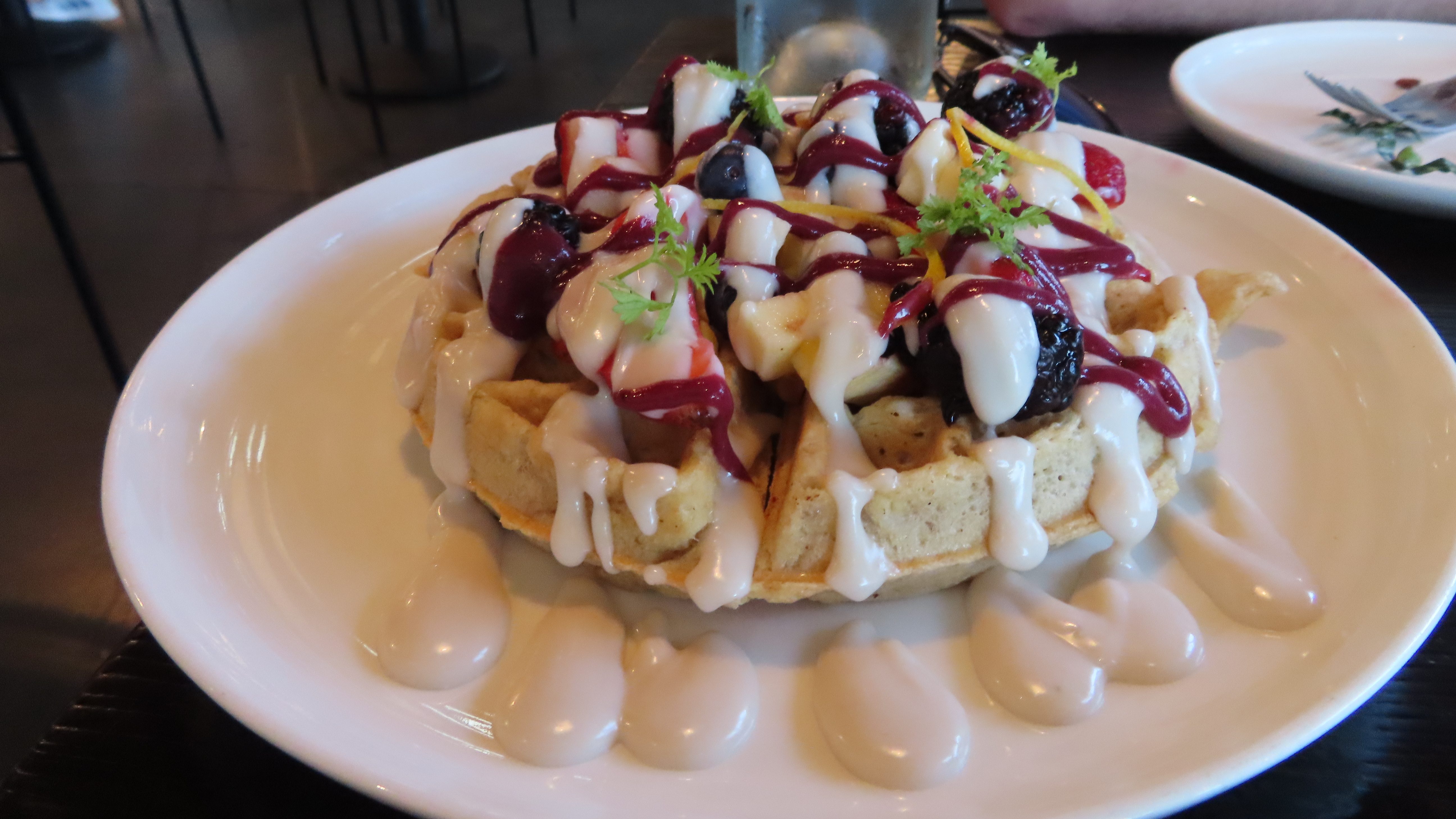 The sloppy waffle with vanilla cream and berry sauce. There are blackberries, blueberries and strawberries. The waffle made from regular and oat flour sits on a white plate.