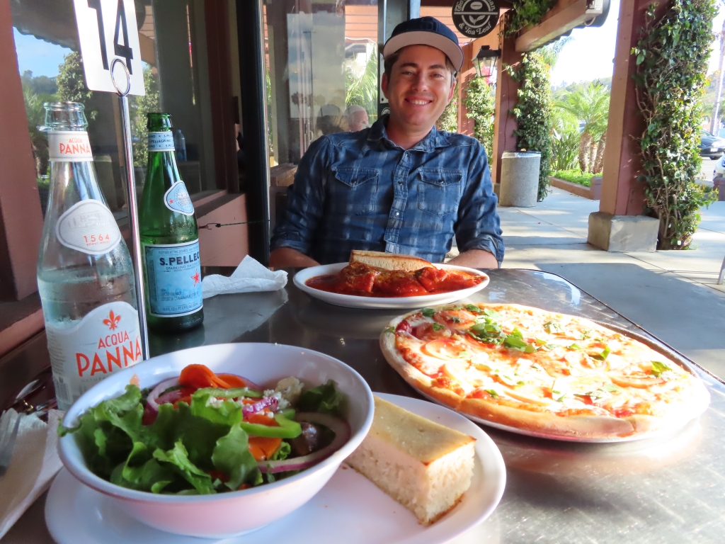 Matthew outside Spirito's smiling in background. In front of him is the table with a margherita pizza, ravioli with country bread and house salad with a slice of bread. There is an acqua panna and a san pellegrino with the number for the order.