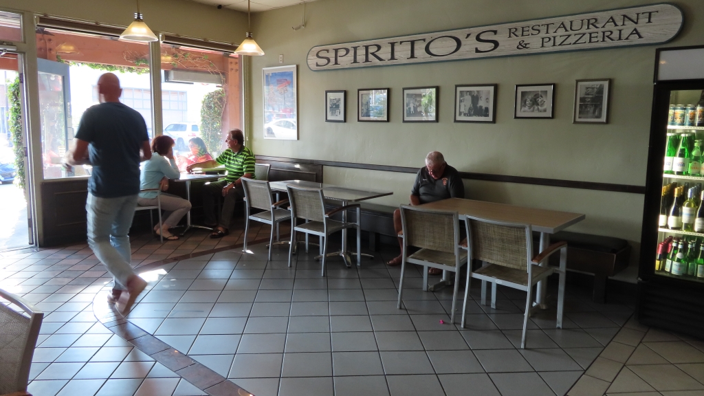 Spirito's dining room has chairs on either end is large space in the middle for customers to walk in and out for take out orders. There is a long sign that reads "Spirito's restaurant and pizza" with photos of their family below. A man looks out the window enjoying a summer afternoon.