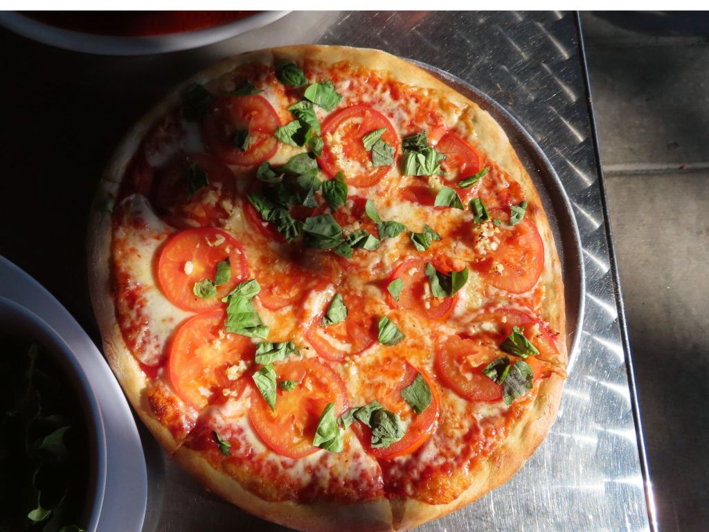 On the table is a margherita pizza served jersey-style. A margherita is a classic chese pizza with whole tomato slices baked in and basil on top. The crust is cooked nicely to be crisp, but not to burn. Shadows from the sun float over the pizza.