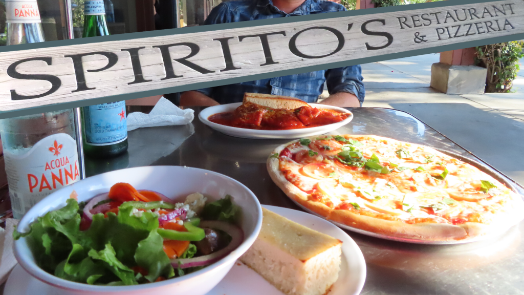 Spirito's Restaurant & Pizzeria sign overlays a photo of a man sitting at an outdoor table at the restaurant. In front of them is a plate of homemade ravioli, pizza, and a bowl of fresh salad with bread. To the side are a bottle of Acqua Panna water and a bottle of San Pellegrino.