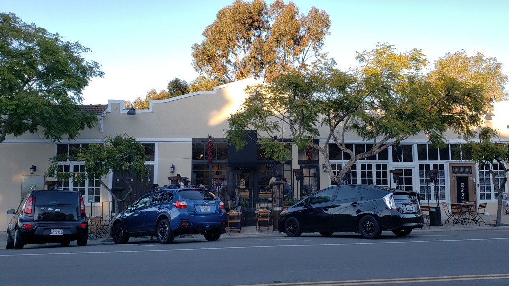 The entrance to the restaurant Wolf in the Woods in the Mission Hills neighborhood of San Diego, CA. Outside is a beige building with a white border on the edge of the roof. The restaurant has an outdoor patio area and a black metal storefront with large windows people can view from the street. There are cars parked out front.
