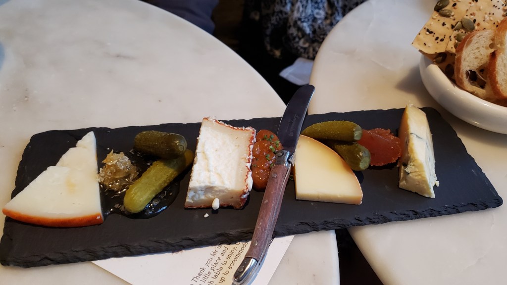 On our two white marble tables is a cheese board on a black marble/charcoal slab with a cup beside it with small slices of baguette and cracker bread. The cheese board has pickles, apricot chutney, soft and hard cheeses with rich flavor notes. They are chosen on a seasonal basis.
