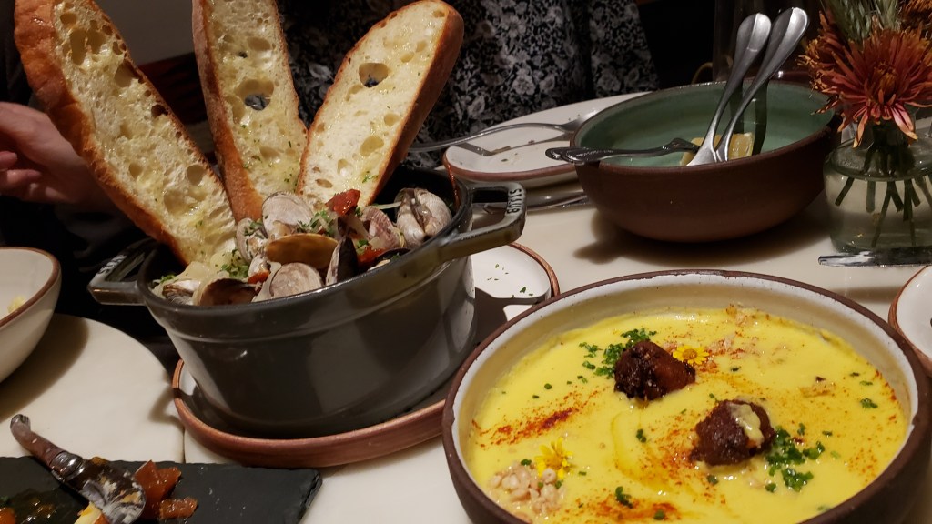 On our table are French clams for two and Corn Pinon soup. The French clams are in a black Staub pot as they were cooked with fennel, onion and garlic in white wine. They're garnished with parsley and shaved chorizo. On the side are three slices of bread spread with farm butter. The sweet corn and pinon soup is served with wildflowers, hatch chile corn fritter, hatch chile dust and crushed pine nuts presented artfully.