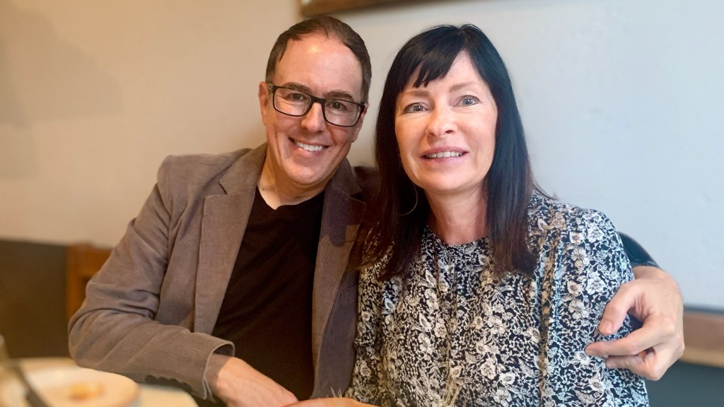 A picture of Heather Sweeney and Kurt Lindemann smiling. Heather is wearing a black and white tulip flower dress. Kurt is in glasses wearing a black t-shirt and light brown, suede blazer.